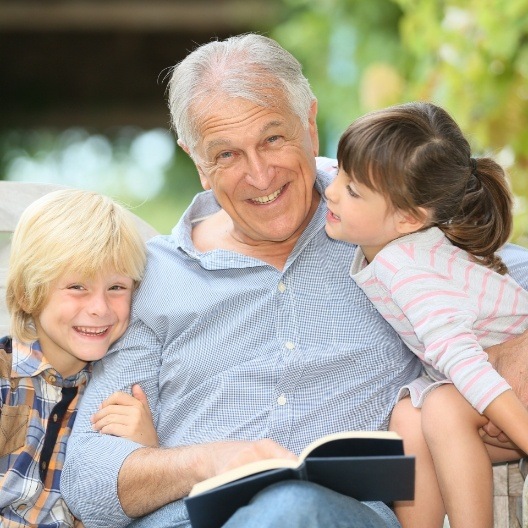 Older man with dental implant supported replacement teeth reading book to his grandkids